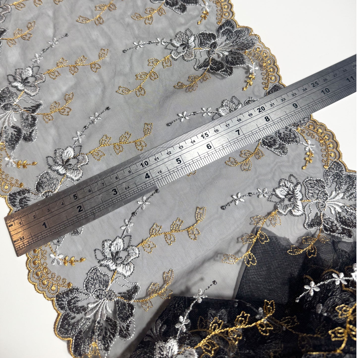 A close-up of a sheer embroidered fabric with floral patterns in black, gray, and gold, shown with a metal ruler for scale on a white background.