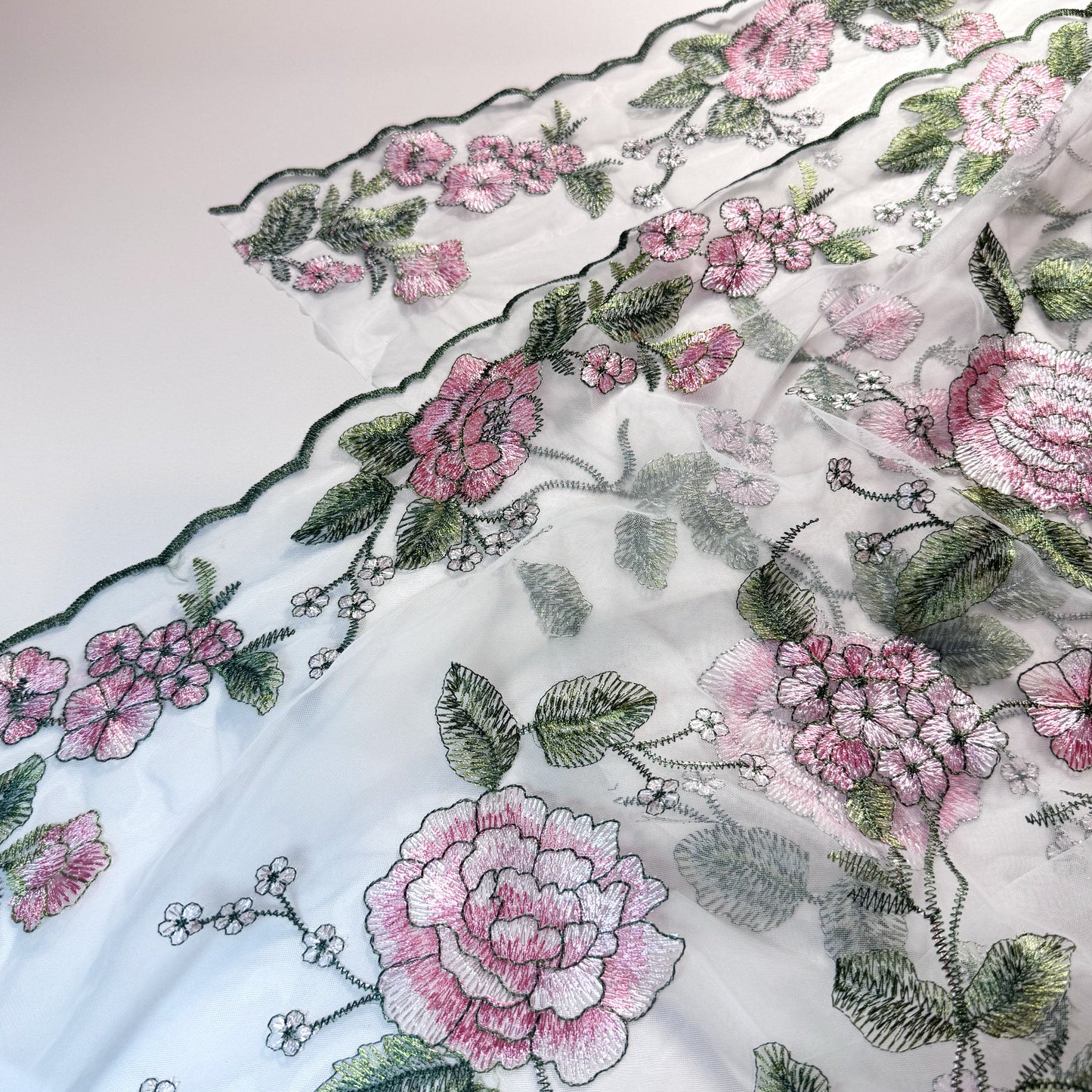 white tulle fabric with green scalloped edges. There are large pink flowers, small white flowers and green leaves all embroidered on the fabric.