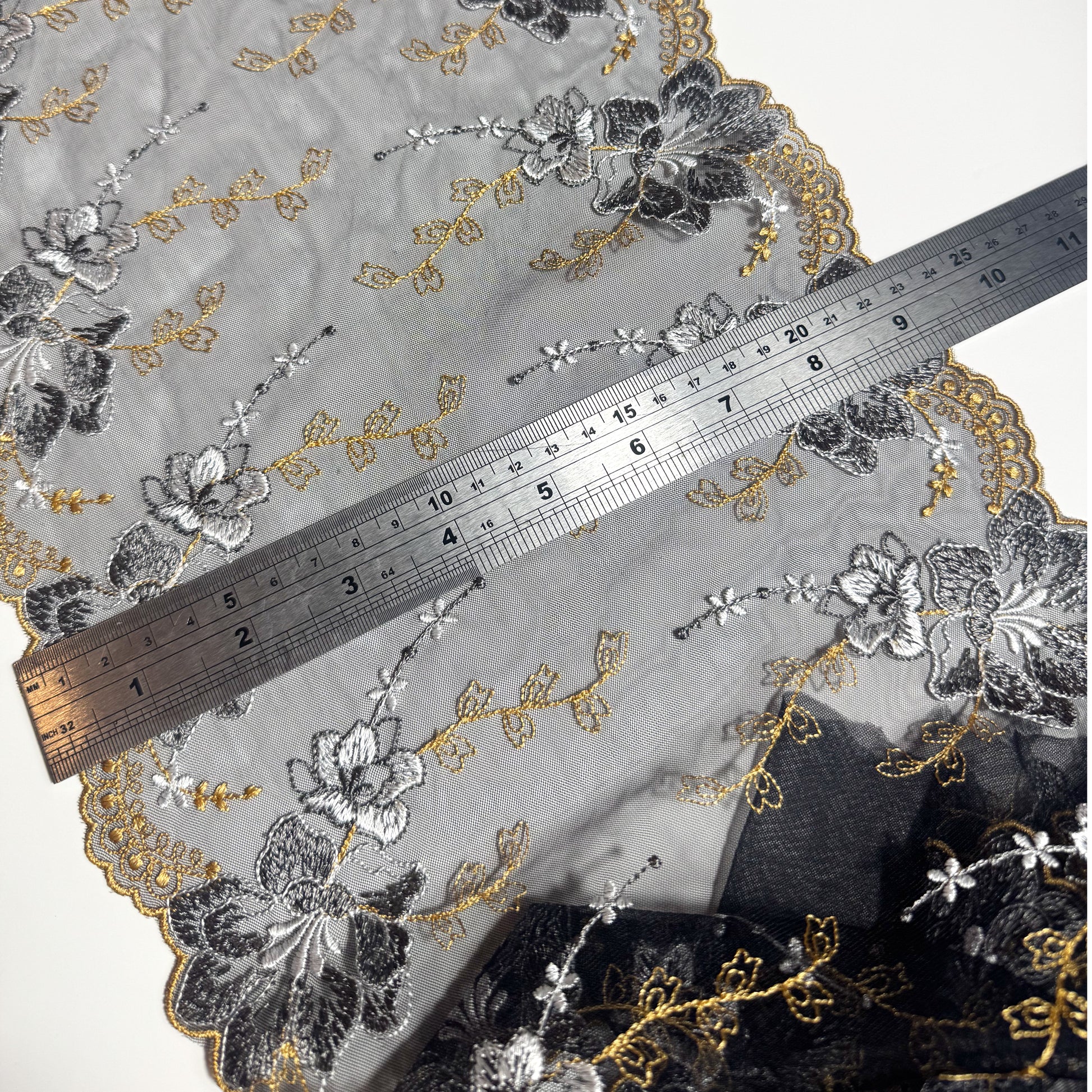A close-up of a sheer embroidered fabric with floral patterns in black, gray, and gold, shown with a metal ruler for scale on a white background.