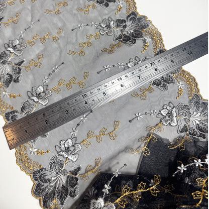 A close-up of a sheer embroidered fabric with floral patterns in black, gray, and gold, shown with a metal ruler for scale on a white background.