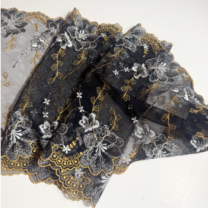 Black mesh lace fabric with white floral embroidery and gold scalloped trim, folded on a white background.